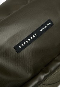 Zielony wodoodporny plecak z bliska, z wyraźną czarną metką z napisem "SUPERDRY" w białych literach. Teksturowane wykończenie.