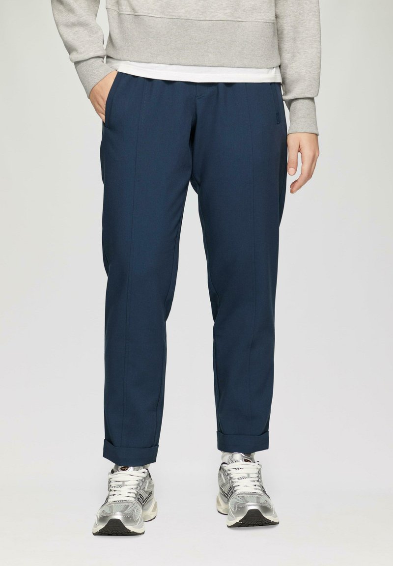 Marineblauwe broek met een taps toelopend ontwerp, gemaakt van een soepele stof, met voorplooien en omgeslagen benen. Gecombineerd met zilverkleurige sport schoenen.