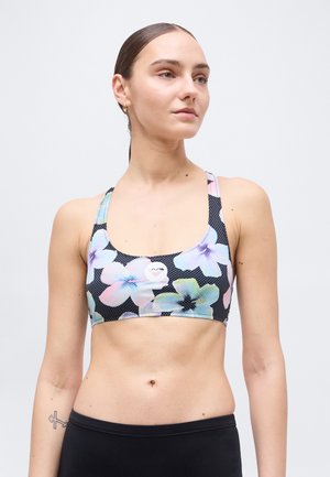 Femme aux cheveux attachés, portant un brassard de sport fleuri et un legging d'entraînement noir, debout devant un fond clair uni.