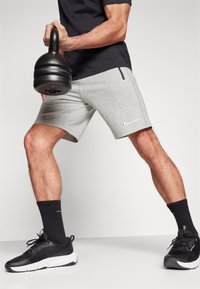 Pantalones cortos deportivos grises con un zipper lateral, que cuentan con un pequeño logo de Champion. El modelo sostiene una kettlebell negra y lleva puestas unas zapatillas deportivas negras y calcetines.
