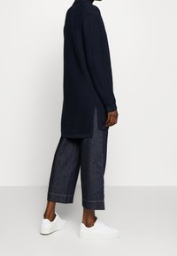 Maglione lungo in maglia blu navy con colletto alto, spacchi laterali e polsini a costine, abbinato a pantaloni cropped in denim scuro larghi e sneakers bianche.