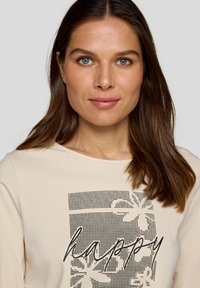 Beige Langarmshirt mit schwarzer Grafik und dem Wort "happy" sowie floralem Muster, aus weichem Baumwollstoff mit glatter Textur.