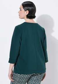 Tee-farbene Bluse mit hohem Kragen, Dreiviertelärmeln und einer geraden Rückennaht, kombiniert mit gemusterten schwarz-weißen Hosen.