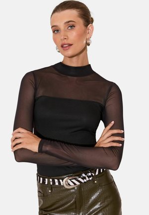 Žena s uhladenými vlasmi ostrihanými dozadu, nosí čierne priehľadné tričko s dlhým rukávom a olivové kožené nohavice s opaskom so zebrovým vzorom, ruky skrížené.