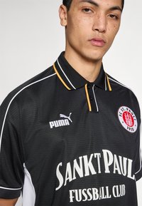Μαύρο πόλο μπλουζάκι με λευκές και κίτρινες λεπτομέρειες. Διαθέτει το λογότυπο της Puma και το κείμενο "Sankt Pauli Fussball Club" σε λευκό, κοντά μανίκια, με διακριτική υφή.
