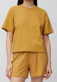 Camiseta de algodón color mostaza con mangas cortas y cuello redondo, que presenta un logo sutil en el pecho y un diseño cropped.