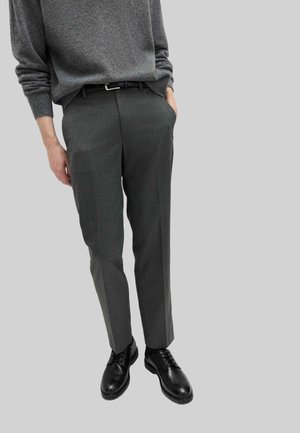 Mann trägt dunkelgraue, maßgeschneiderte Hose, schwarzen Gürtel, schwarze Lederschuhe und einen grauen Pullover mit langen Ärmeln, eine Hand in der Tasche.