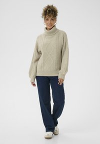 Frau mit lockigem Haar, die einen beigen Strickrollkragenpullover, dunkelblaue Jeans und weiße Sneakers trägt und vor einem weißen Hintergrund steht.