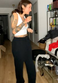 Femme avec des tatouages portant un débardeur blanc et un pantalon noir se tient dans une pièce encombrée avec des vêtements sur un canapé et des objets sur une table en verre.