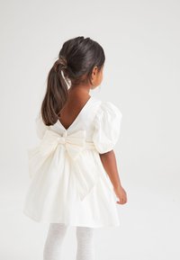 Robes en coton blanc à manches bouffantes, design avec dos en V et grand nœud à la taille. Des collants texturés légers avec des motifs en forme de cœur complètent le look.