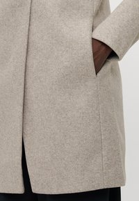 Beige wollen blend jas met een rechte snit, zijzakken en een iets gestructureerde afwerking. Zichtbare naden langs de randen.