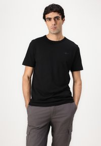 Camiseta de algodón negra con cuello redondo, mangas cortas y un pequeño logo negro en el pecho. Lleva puesta con pantalones cargo grises.