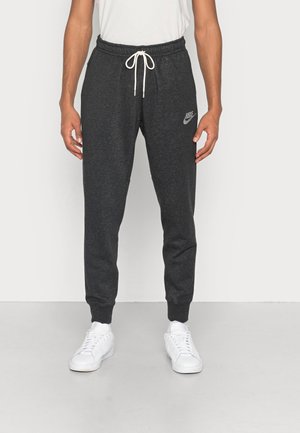 Man draagt donkergrijze Nike joggingbroek met witte trekkoord en witte sneakers, staand tegen een effen lichte achtergrond.