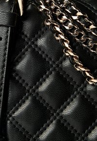 Sac à main matelassé noir avec extérieur synthétique texturé, doté d'une bandoulière à chaîne dorée et de détails assortis, fermé par une fermeture éclair noire.