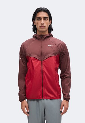 Muž s dlhými tmavými vlasmi, ktorý má na sebe bordovo-červenú bundu Nike so zipsom a kapucňou a sivé nohavice, stojaci pred jednoliatym pozadím.
