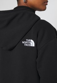 Osoba nosící černou mikinu s kapucí s bílým vyšitým logem The North Face na horní části zad u ramene.