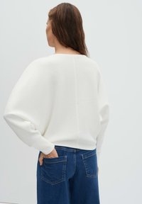 Vrouw met lang bruin haar, gekleed in een witte oversized trui en blauwe spijkerbroek, staand met één hand in haar achterzak.