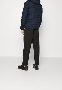 Mann in schwarzer Daunenjacke, lockeren schwarzen Hosen und schwarzen Sneakers, der vor einem schlichten, hellen Hintergrund steht und sich abwendet.