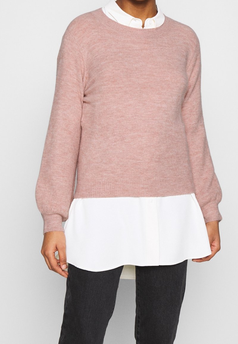 Jasnoróżowy dziergany sweter z prążkowanym obrzeżem i bufkami na rękawach, noszony na białej koszuli z kołnierzykiem, zestawiony z ciemnymi dżinsami.