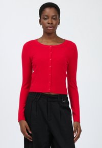 Cardigan rosso lavorato a maglia con scollo rotondo, dotato di una fila di sei bottoni, maniche lunghe e orlo a costine. Abbinato a pantaloni neri.