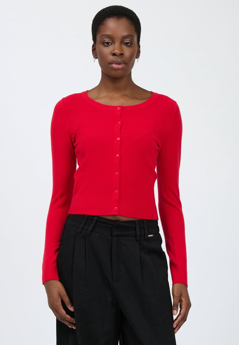 Cardigan rosso lavorato a maglia con scollo rotondo, dotato di una fila di sei bottoni, maniche lunghe e orlo a costine. Abbinato a pantaloni neri.