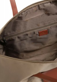 Intérieur d'un sac tote beige avec une texture lisse, comportant une poche zippée marron et un logo embossé sur une pièce en cuir.