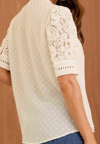 Blusa bianca in tessuto testurizzato con pois, maniche a sbuffo in pizzo e dettagli decorativi lungo l'orlo e i polsini.