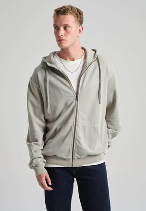 Junger Mann mit lockigem Haar, der einen hellgrauen Zip-Hoodie und ein weißes Shirt trägt, schaut nach links mit den Händen in den Taschen vor schlichtem Hintergrund.