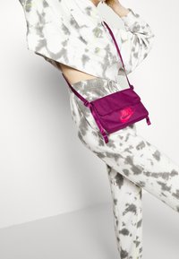 Lila tygaxelväska med lock, med en synlig Nike-logotyp i rosa. Väskan har justerbara remmar och en slät yta.