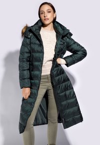 Long, dark green puffer coat with a fur-trimmed hood, zip closure, and quilted design. Worn over a light sweater and olive green pants.