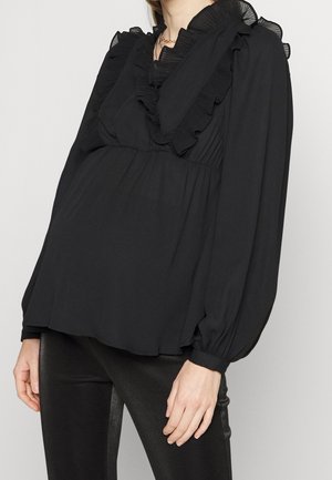 Femme portant une blouse noire à manches longues avec col et épaules à volants, associée à un pantalon noir ajusté, sur un fond uni.