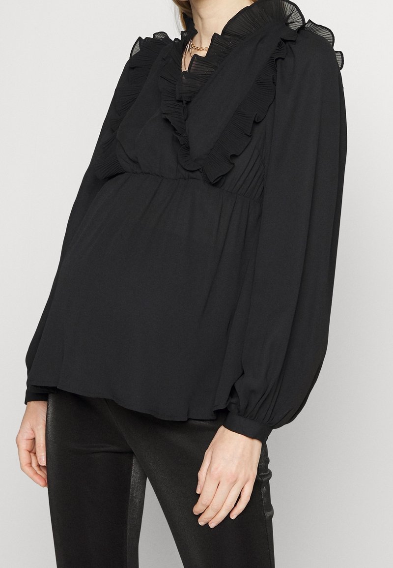 Femme portant une blouse noire à manches longues avec col et épaules à volants, associée à un pantalon noir ajusté, sur un fond uni.