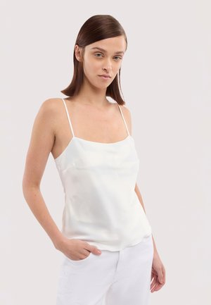 Jeune femme aux cheveux bruns et raides portant un caraco en satin blanc à fines bretelles et un pantalon blanc, debout devant un fond clair.