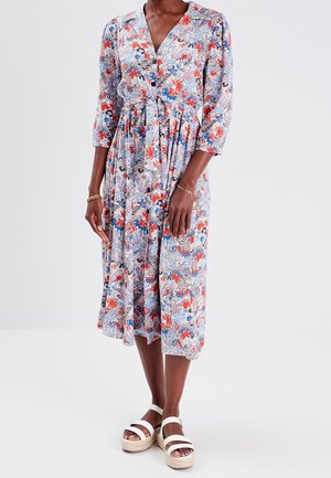 Femme portant une robe fleurie mi-longue à manches trois-quarts et des sandales blanches à plateforme, debout sur un fond uni.