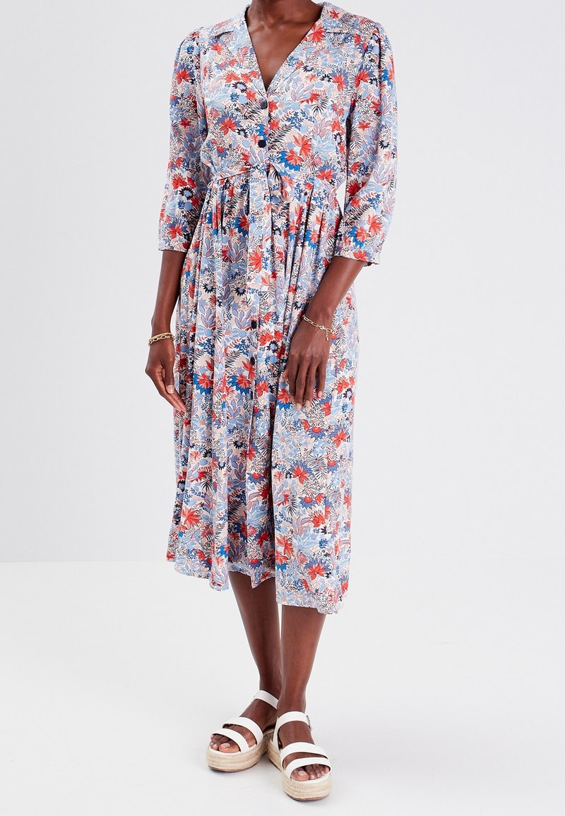Femme portant une robe fleurie mi-longue à manches trois-quarts et des sandales blanches à plateforme, debout sur un fond uni.