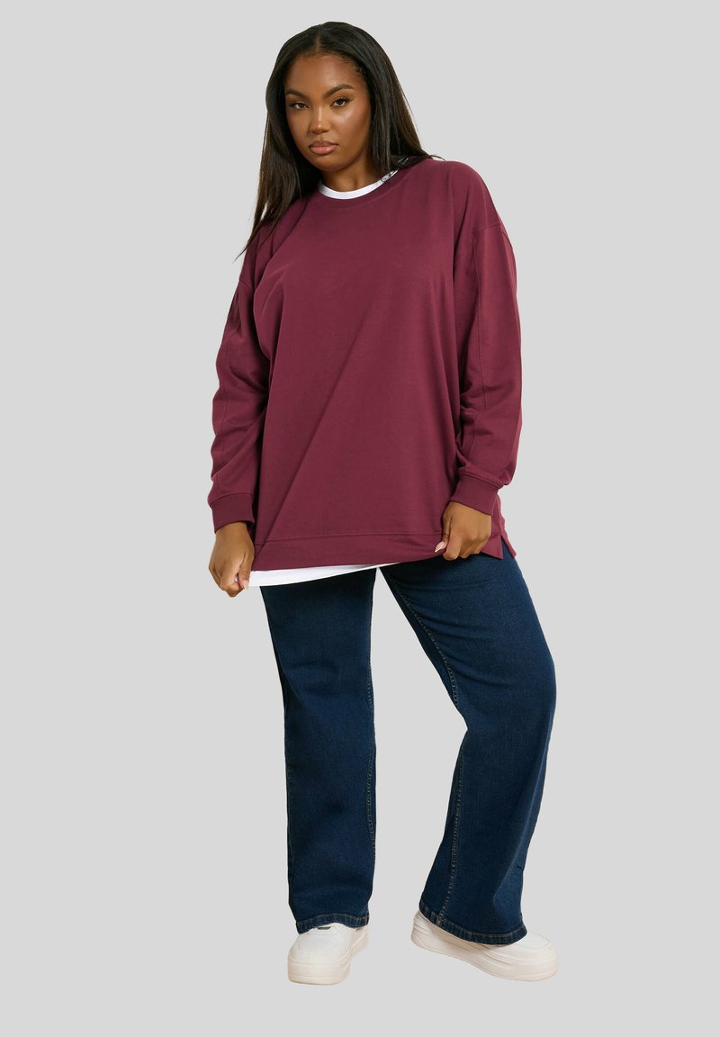 Nainen pukeutuneena löysään marooniin collegepaitaan, jonka päällä on valkoinen paita, tummansiniset farkut ja valkoiset tenniskenkä, seisoo yksinkertaista taustaa vasten.