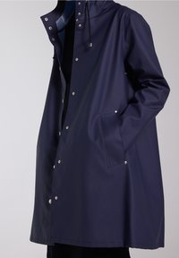 Tmavě modrý plášť proti dešti s kapucí, vyrobený z nepromokavého materiálu. Má patentní knoflíky, přední kapsu a stahovací šňůrku na kapuci.
