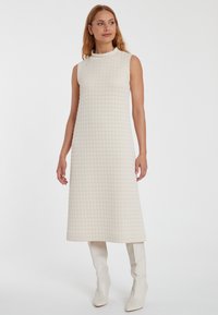 Strick ärmelloses Kleid mit hohem Kragen, das ein strukturiertes, gesteppteres Muster in Creme zeigt. Kombiniert mit kniehohen weißen Stiefeln.