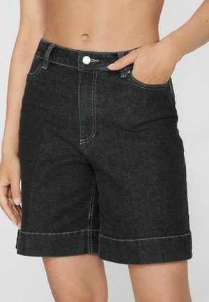 Kobieta ubrana w czarne szorty jeansowe z białym przeszyciem, guzik z przodu, szlufki na pasek i jedną ręką w kieszeni na jednolitym tle.