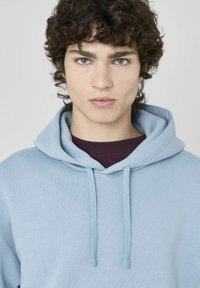 Haut à capuche bleu clair avec cordon de serrage, doté d'un col côtelé bordeaux et d'une texture douce. Le modèle a des cheveux bouclés et une expression neutre.