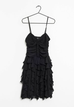 Robe noire sans manches avec corsage froncé et jupe à volants superposés suspendue à un cintre en fil métallique fin sur fond blanc.
