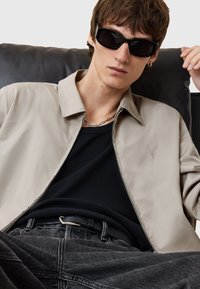 Lichtbeige jas met een kraagontwerp, gedragen over een zwarte getailleerde shirt. Donkergrijze denim jeans en zwarte zonnebril, minimale accessoires zichtbaar.