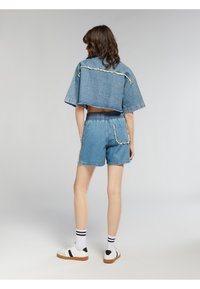 Giacca denim cropped con bordi sfrangiati, abbinata a pantaloni shorts in denim con vita elastica e tasca laterale. Indossata con sneakers bianche e calzini a righe.