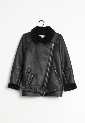 Veste en cuir noire avec col et poignets en fourrure, fermeture éclair asymétrique à l’avant, trois poches zippées et une ceinture à la taille, suspendue à un crochet.