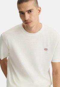 T-shirt bianco in cotone con scollo rotondo e maniche corte. Presenta un piccolo logo rosso sulla parte sinistra del petto. Tessuto morbido.