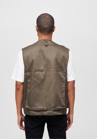 Olivgrüne Weste aus glattem Stoff, mit einer großen Tasche auf der Rückseite und einer Schlaufe am Hals zum Aufhängen. Weißes T-Shirt darunter.