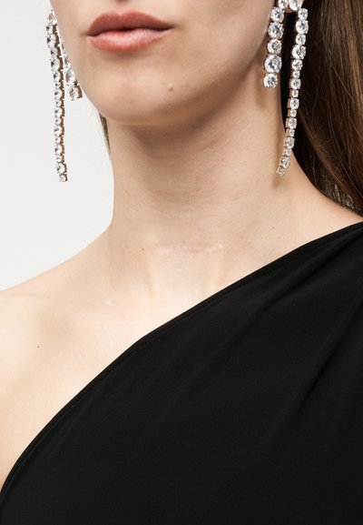 Κοντινό πλάνο μιας γυναίκας που φοράει μακριά κρεμαστά σκουλαρίκια με στρας και μαύρο τοπ με έναν ώμο, εμφανίζοντας τα χείλη και τον λαιμό της.