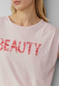 Lichtroze t-shirt met een ronde hals, voorzien van rode, textuurblokletters die "BEAUTY" spellen. Korte mouwen, zachte stof.