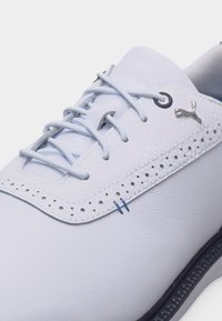 Λευκό γκολφ παπούτσι από δέρμα με ανοιχτό μπλε κορδόνια, διακοσμητικές τρύπες και ασημένιο λογότυπο Puma κοντά στα κορδόνια.