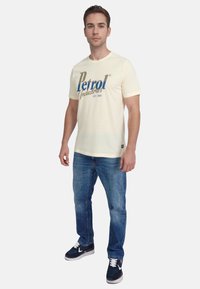 Cremefarvet bomulds t-shirt med blå og guld "Petrol Industries" tekst, parret med lyseblå jeans og marineblå sneakers.